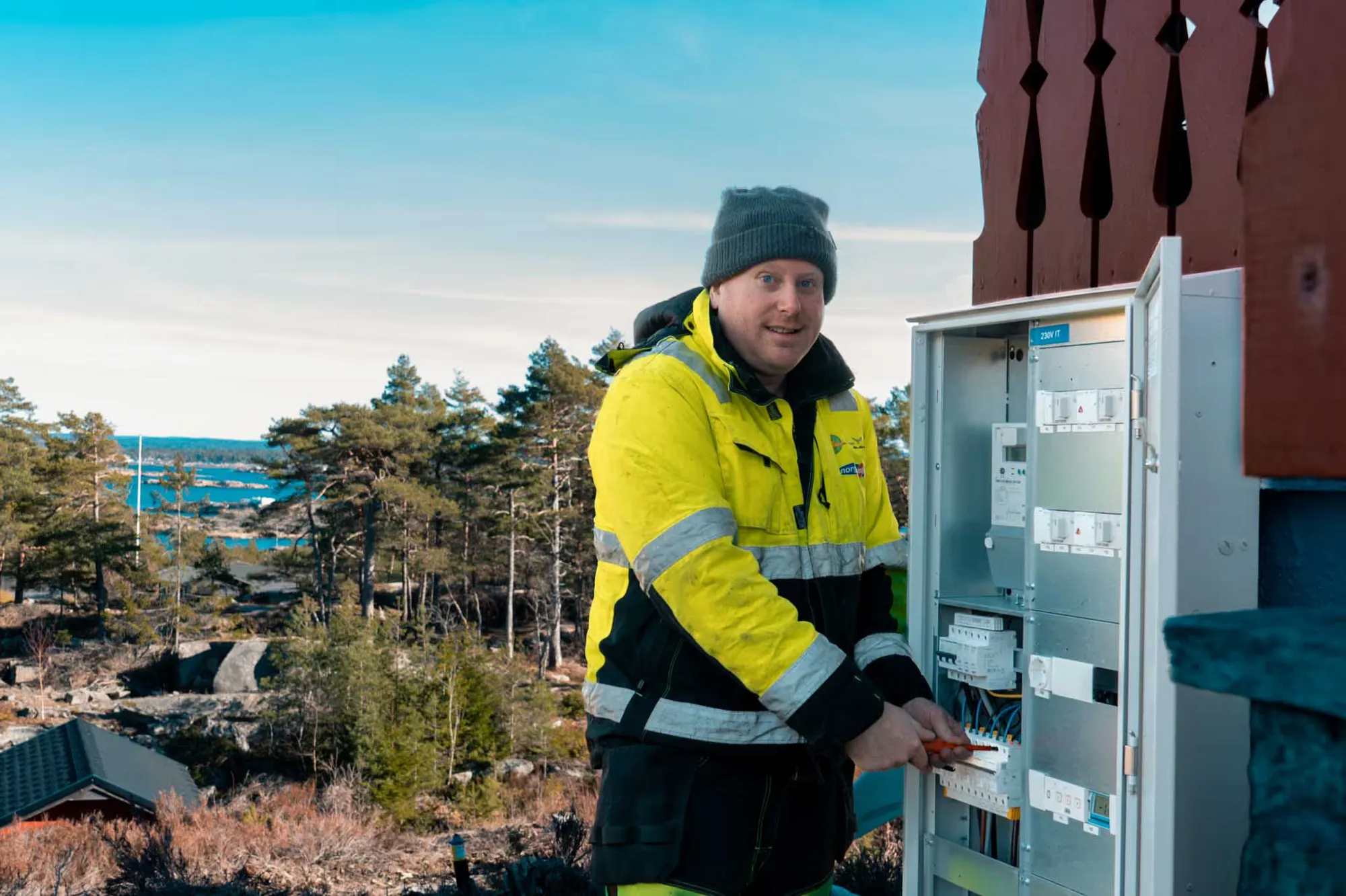 Elektriker fra LYN Elektro i refleksjakke ved et åpent el-skap, med sjøen og svaberg bak